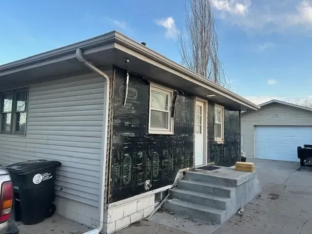 House with damaged siding and exposed black underlayment. Gray siding, concrete steps, and blue sky.