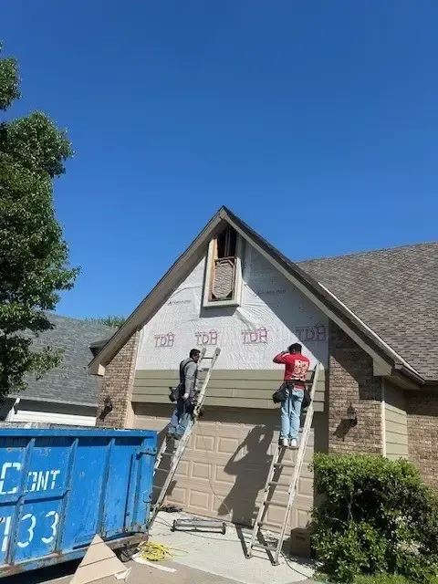 Two workers on ladders installing siding on a house with a blue sky. A dumpster is on the left.