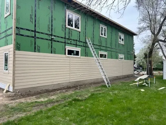 Building exterior under construction with beige siding installed on the lower level and green sheathing above. Ladder leans against the building.