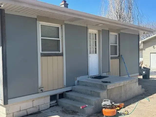 Exterior view of a house with gray siding, beige panels, white door, concrete steps, and a clear blue sky.