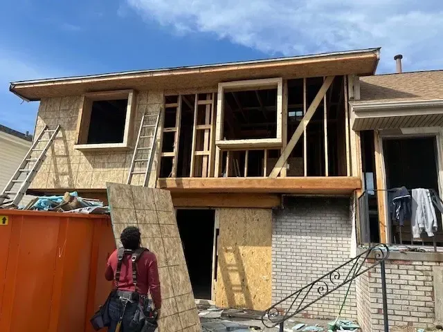 Construction of a house addition; wooden frame, plywood, open windows, worker with plywood, orange dumpster, sunny day.