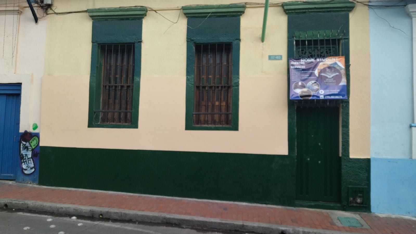 Fachada de edificio con molduras verdes, tres ventanas enrejadas, una puerta y un cartel en una pared beige.