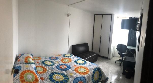 Dormitorio con cama, sillón, escritorio y armario. Paredes blancas, edredón estampado y suelo gris.