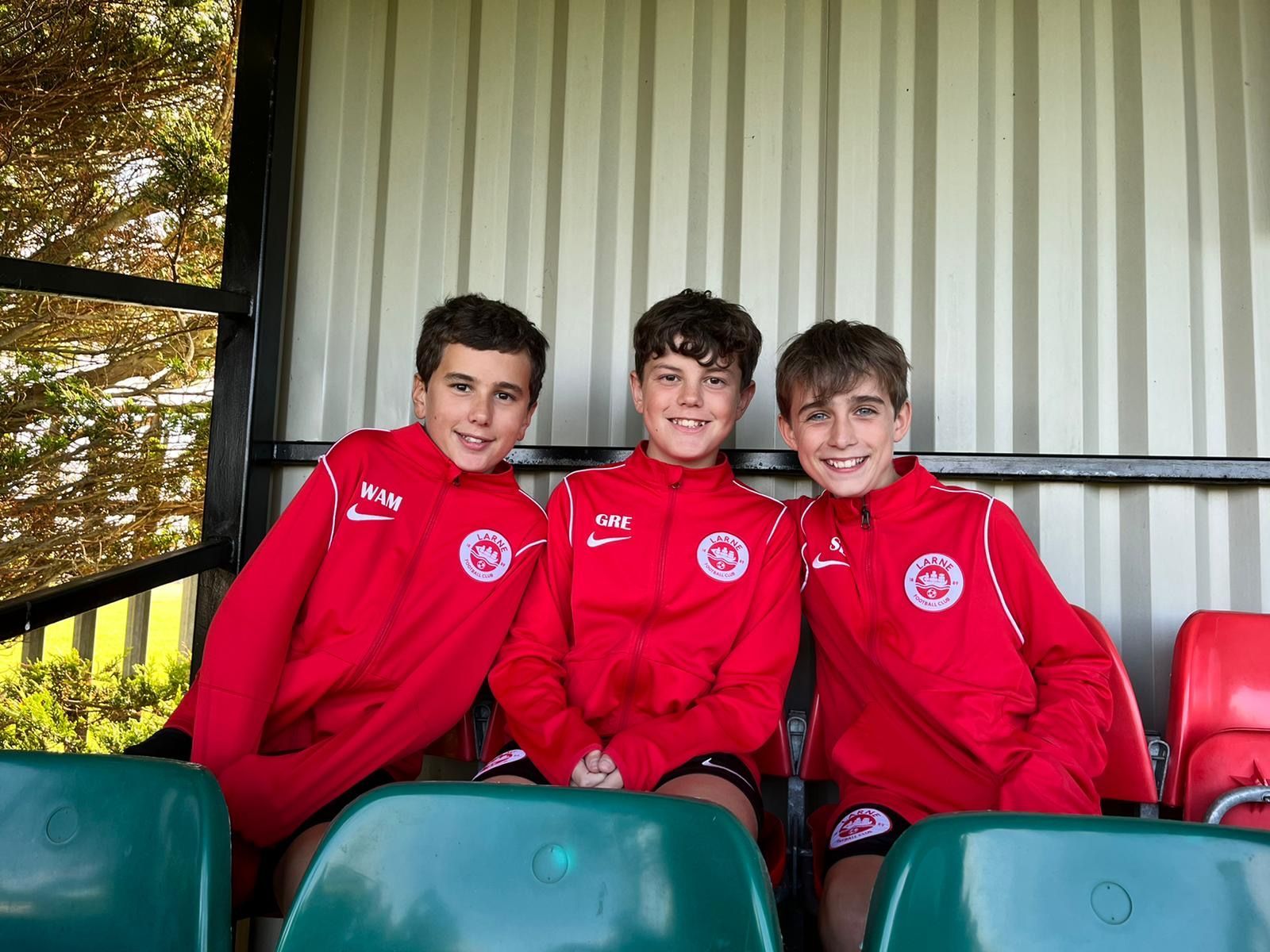 Three young boys in red tracksuits smiling at a sporting event, seated in green and red stadium seating.