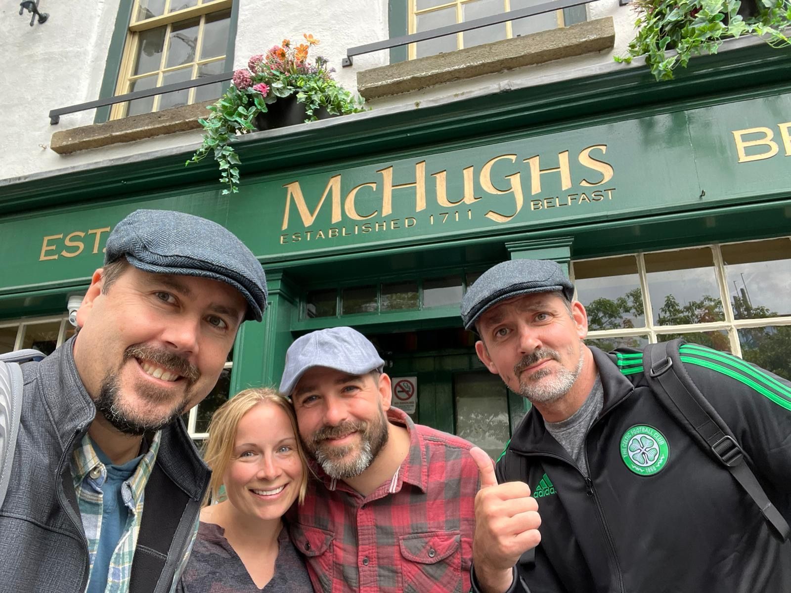 Four people smile in front of McHughs Bar, green sign above door. Two men wear flat caps.