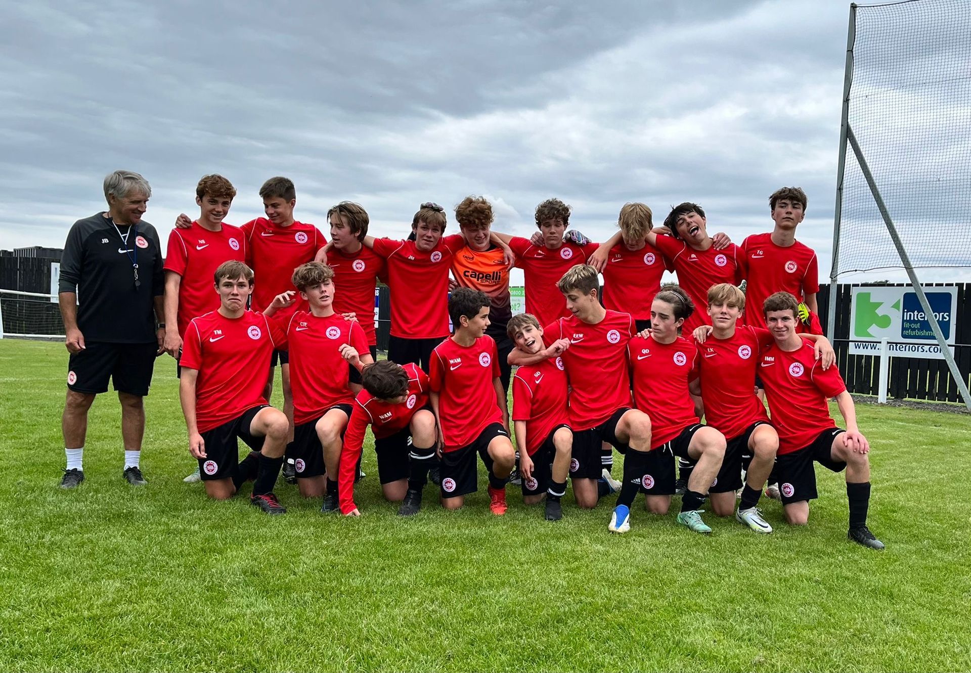 Youth soccer team in red jerseys pose on a green field with their coach, under cloudy skies.