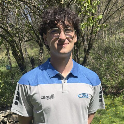 Man with glasses wearing a blue, gray, and white polo shirt, standing in front of trees.