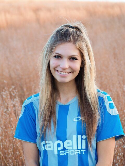 Woman with long blonde hair smiles, wearing a blue soccer jersey, outdoors with tall grass background.