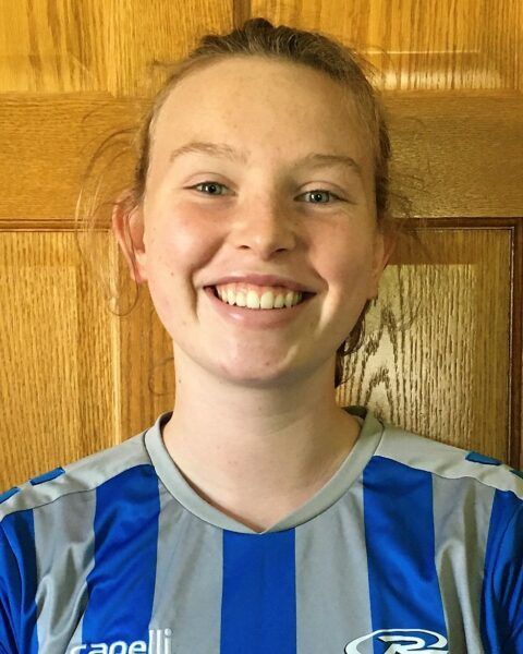 Woman smiling, wearing a blue and gray soccer jersey, in front of a wooden door.