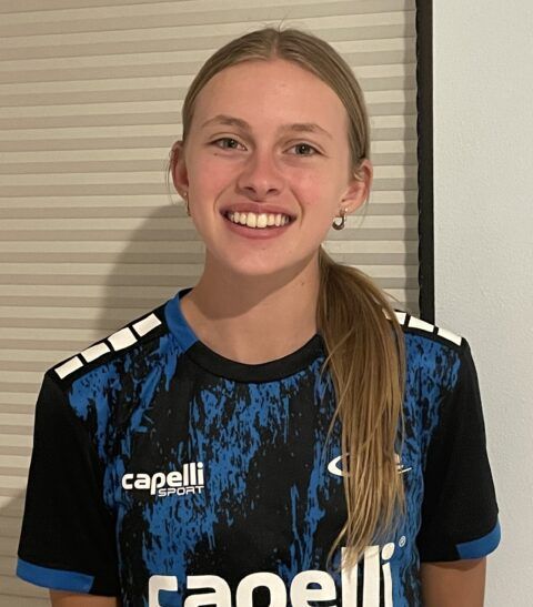 Woman with blonde hair in a blue and black jersey smiles, standing against a neutral background.