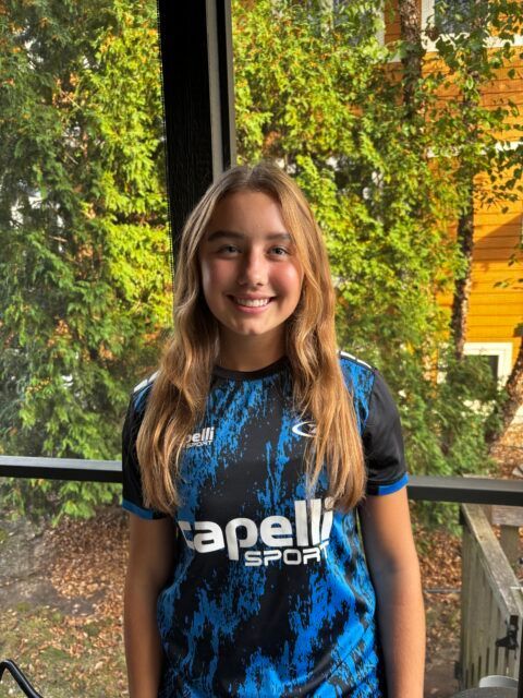 Young person in blue soccer jersey smiles in front of trees.