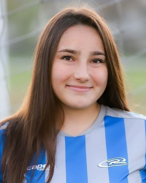 Woman in soccer jersey, smiling at the camera, with long brown hair. Blue and grey striped jersey, blurred background.