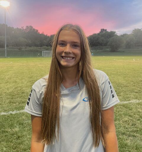 Woman with long blonde hair smiles, wearing a light blue soccer jersey, standing on a green soccer field at dusk.