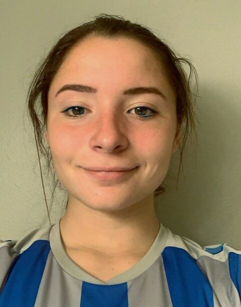 Woman with brown hair smiling, wearing a blue and gray striped shirt, neutral background.