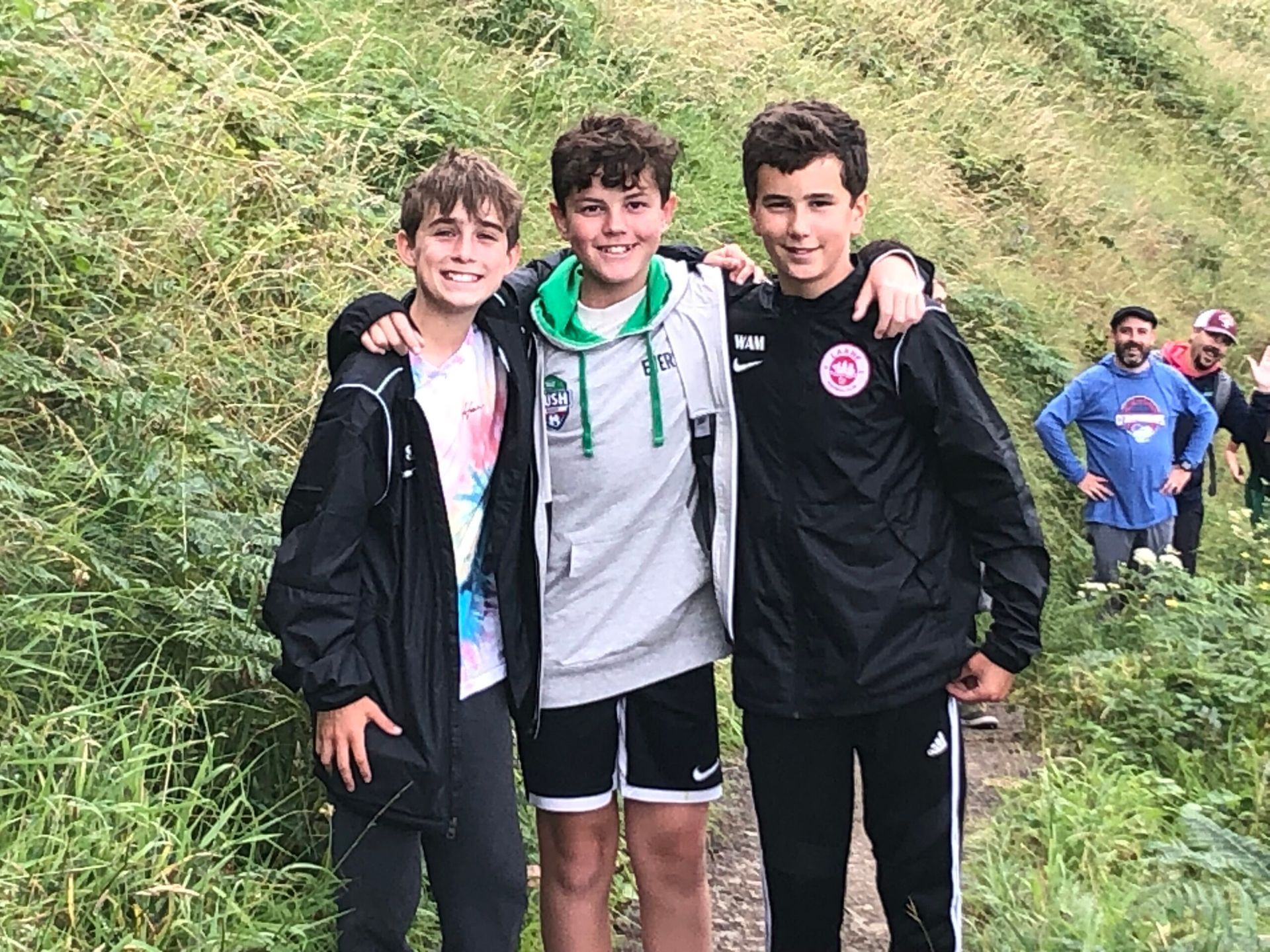 Three boys standing closely together on a trail, smiling. One has arms around shoulders. Green vegetation in background.