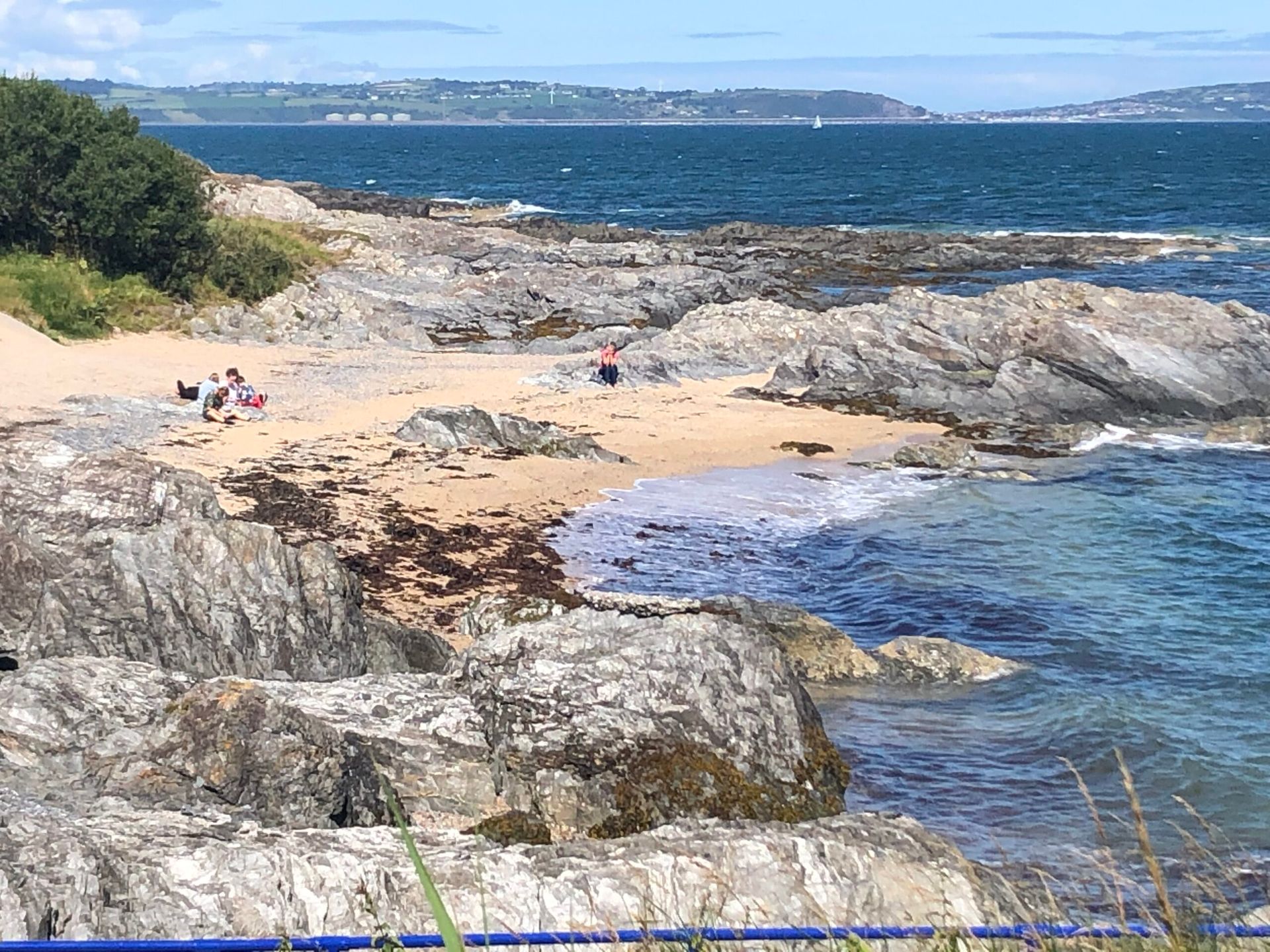 Rocky beach with small sandy cove. People on the beach, blue sea, and green vegetation in the background.