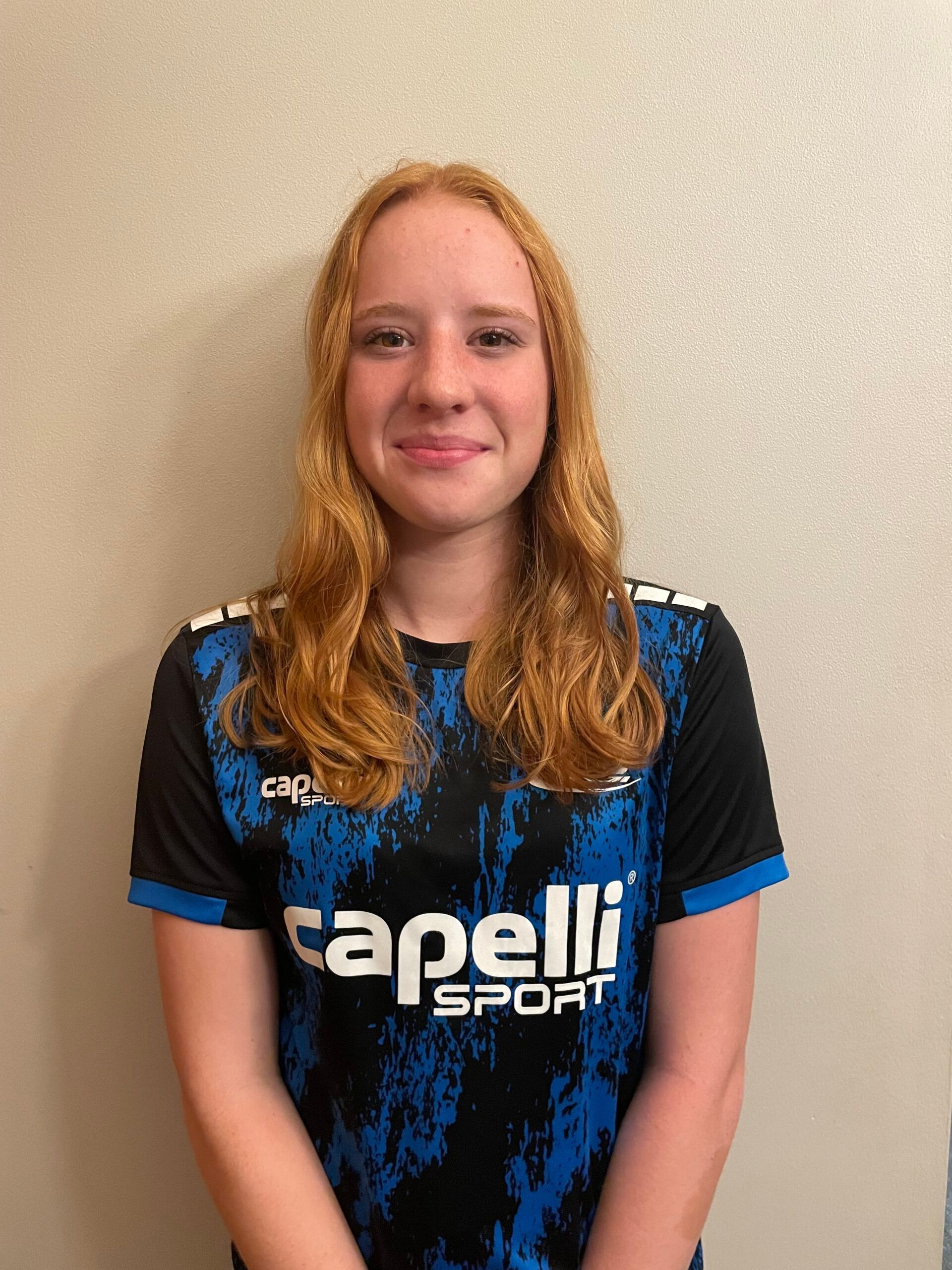 Woman with reddish hair in a blue and black Capelli Sport shirt smiles.
