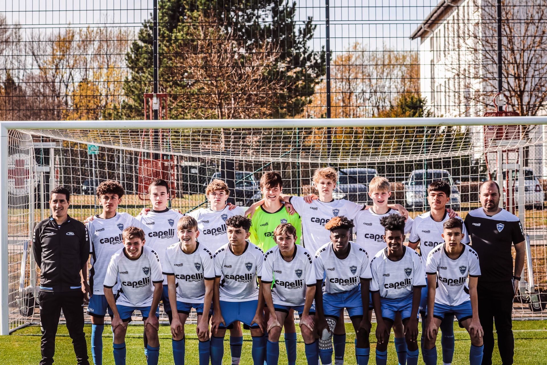 Soccer team posing in front of goal; players in white and blue uniforms with coaches.