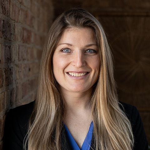 Woman with long blonde hair smiles, wearing a black blazer and blue shirt, brick wall background.