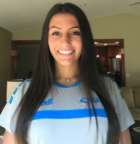 Woman with long dark hair, wearing a light blue sports jersey, smiles at the camera. Interior setting.