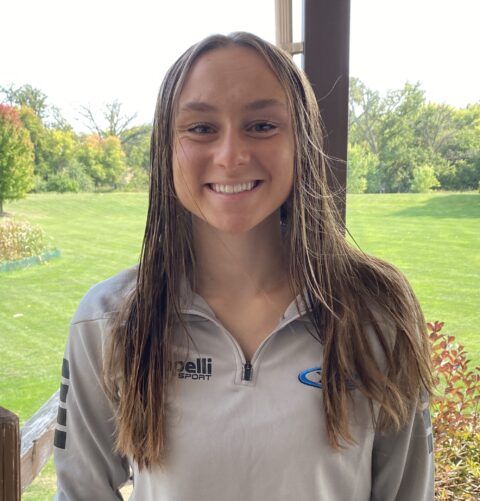 Woman with long brown hair smiling, wearing a gray sweatshirt, standing outdoors.