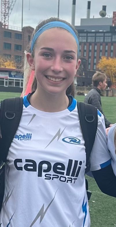 Girl in white soccer jersey with blue accents, smiling outdoors.