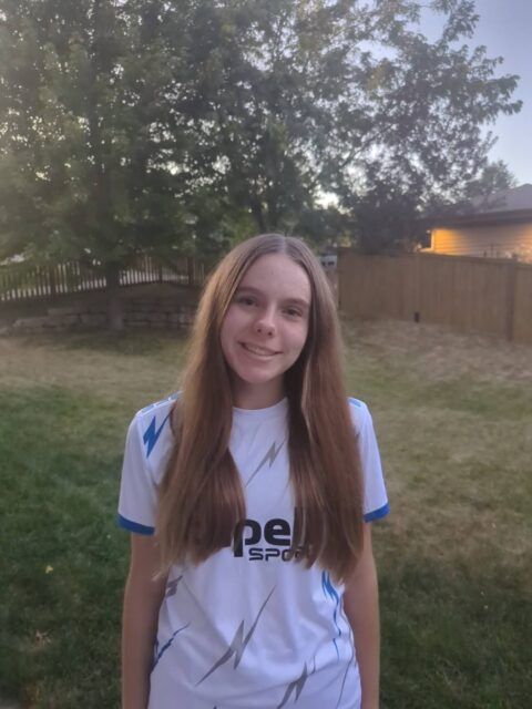Young person with long brown hair smiling outdoors, wearing a white sports jersey.