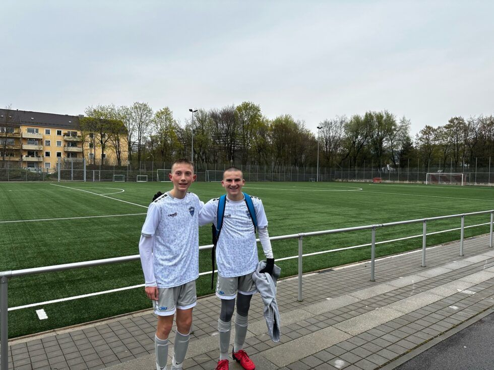 Two soccer players in white jerseys stand on a walkway beside a green field; overcast sky.