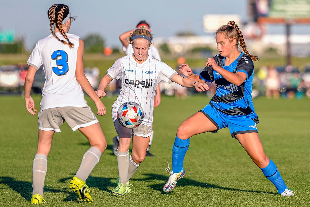 Three soccer players on a field: one with the ball, flanked by two others, one in blue and one in white.