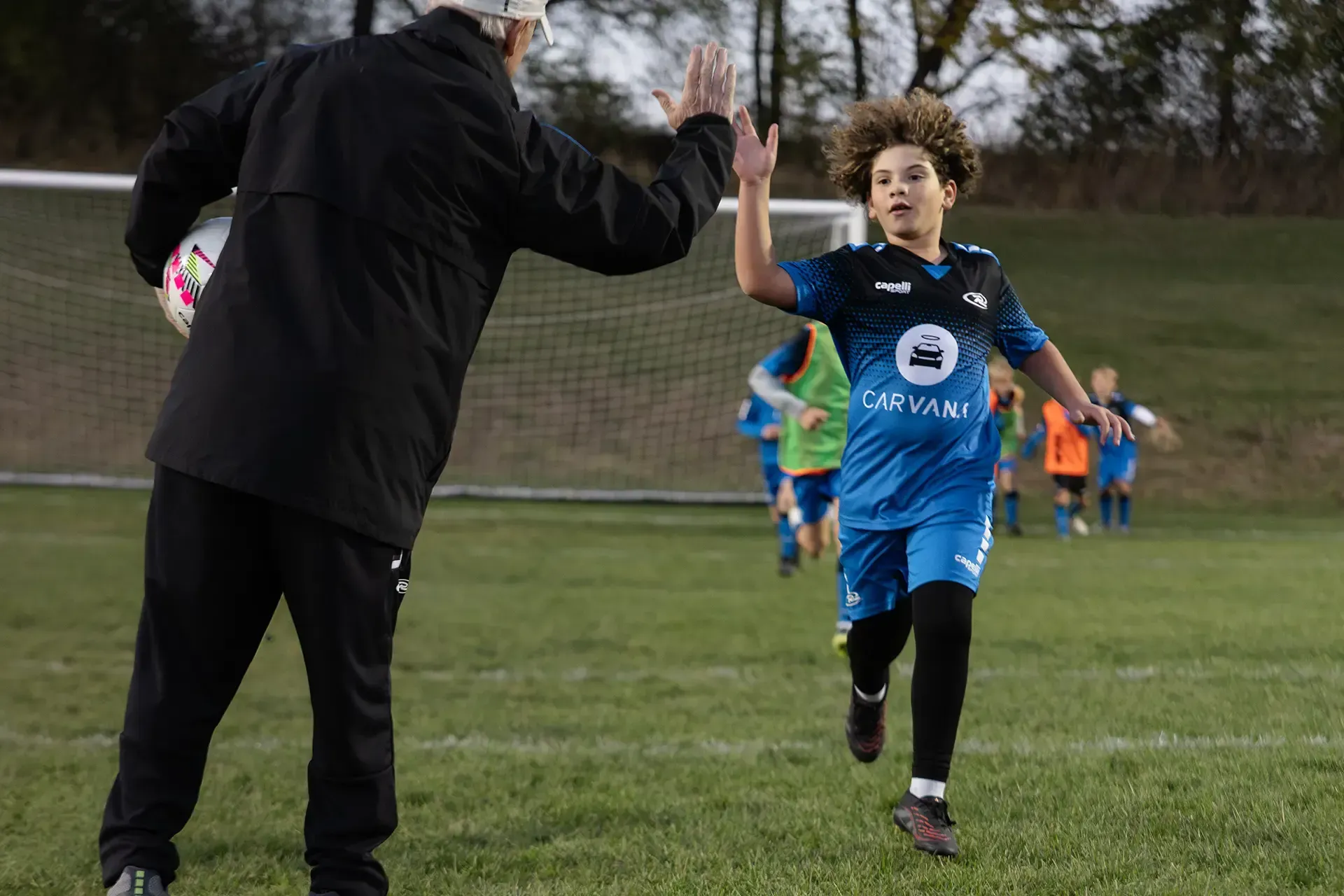 Three soccer players on a field: one with the ball, flanked by two others, one in blue and one in white.