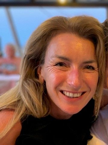 Lachende vrouw met lang blond haar en een zwarte top, die binnen dicht bij de camera leunt.