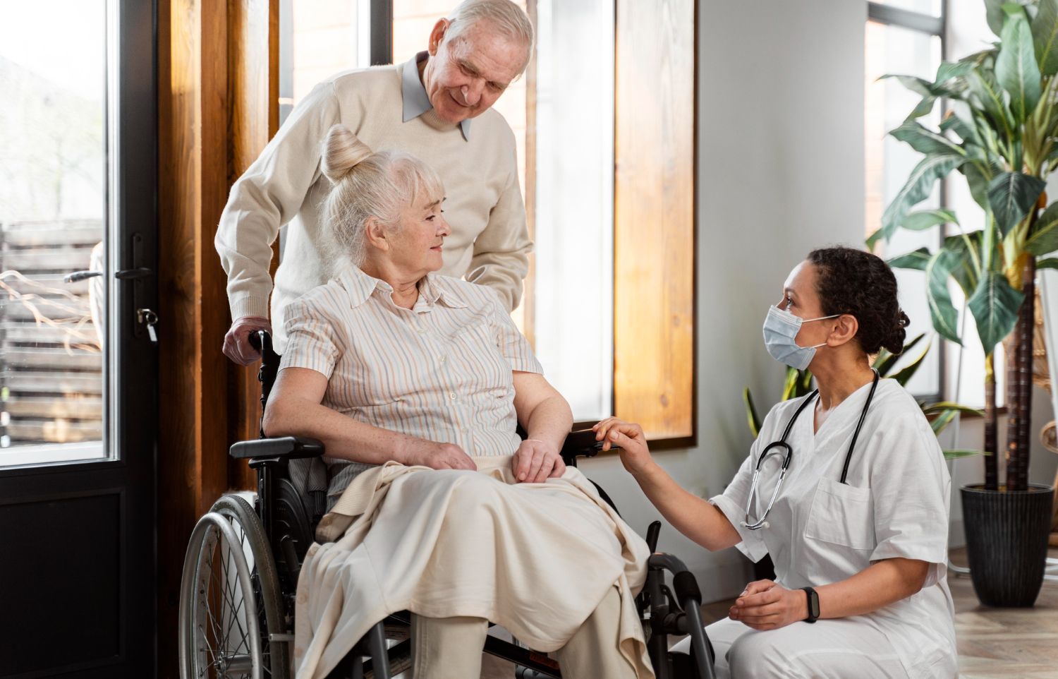 Elderly woman in wheelchair with husband and nurse, indoors. Nurse in mask touches woman's hand.