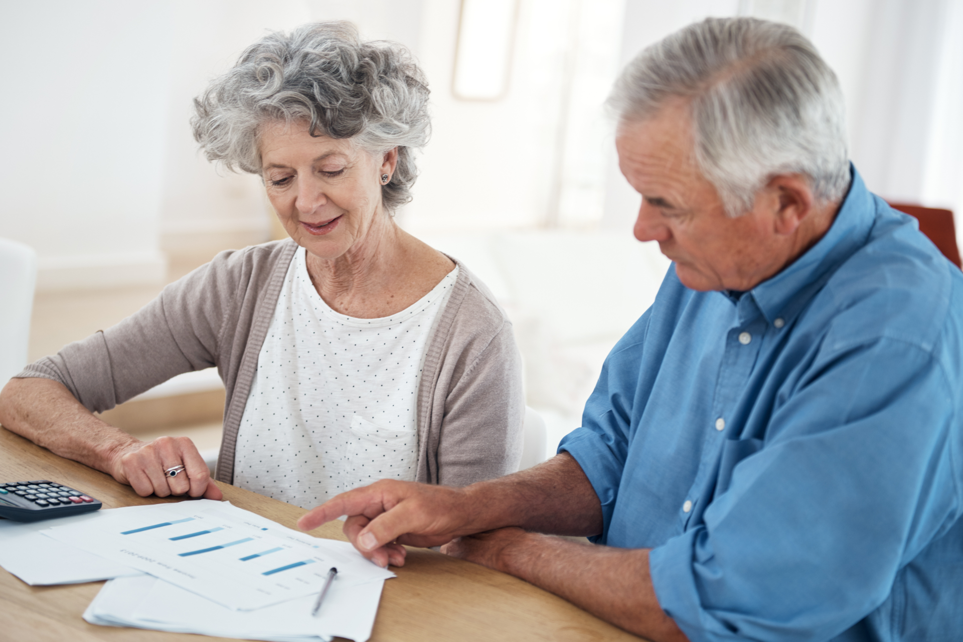 Elderly couple reviews financial documents at a table; man points, woman looks on.