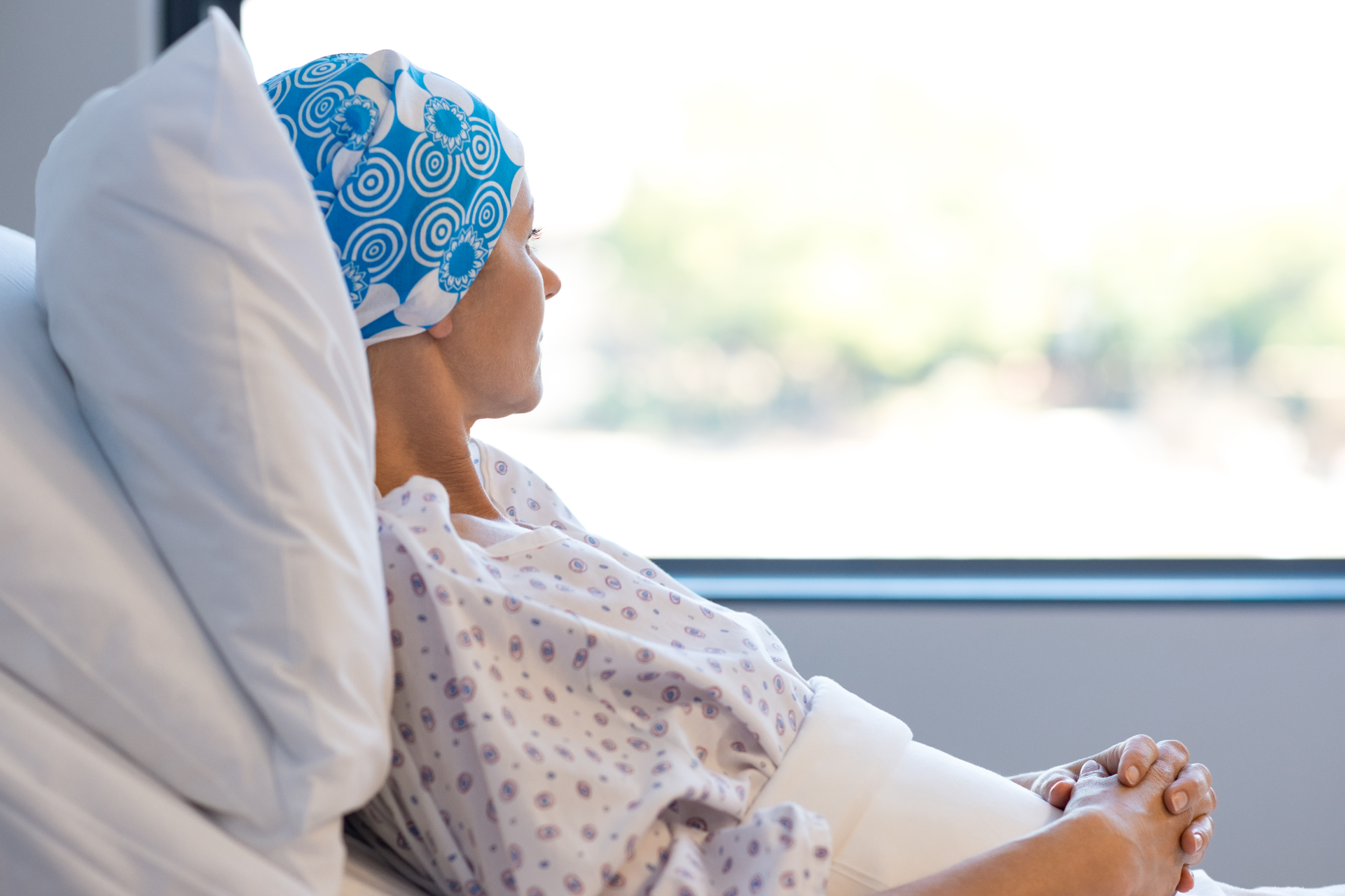 Woman in hospital bed wearing headscarf, looking out window, hands clasped.