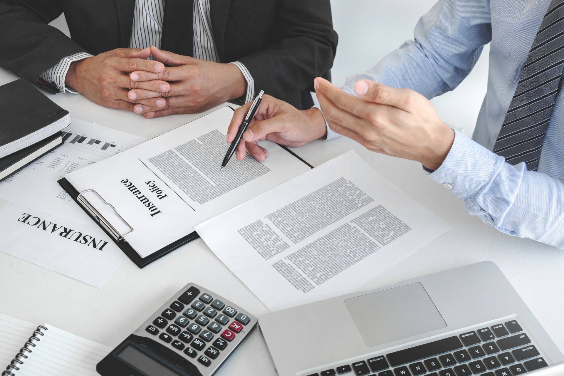 Two people review insurance documents at a desk with a calculator and laptop.