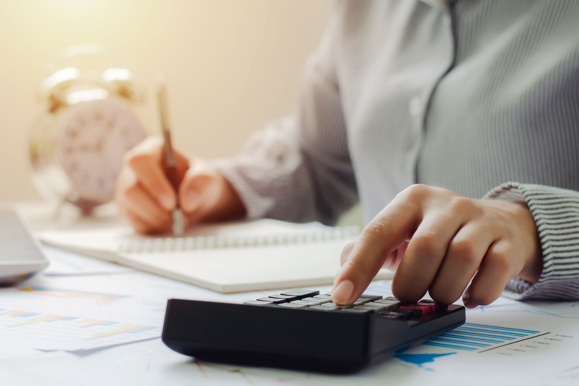 Person calculating on a calculator, writing notes with a pen, surrounded by papers and an alarm clock.
