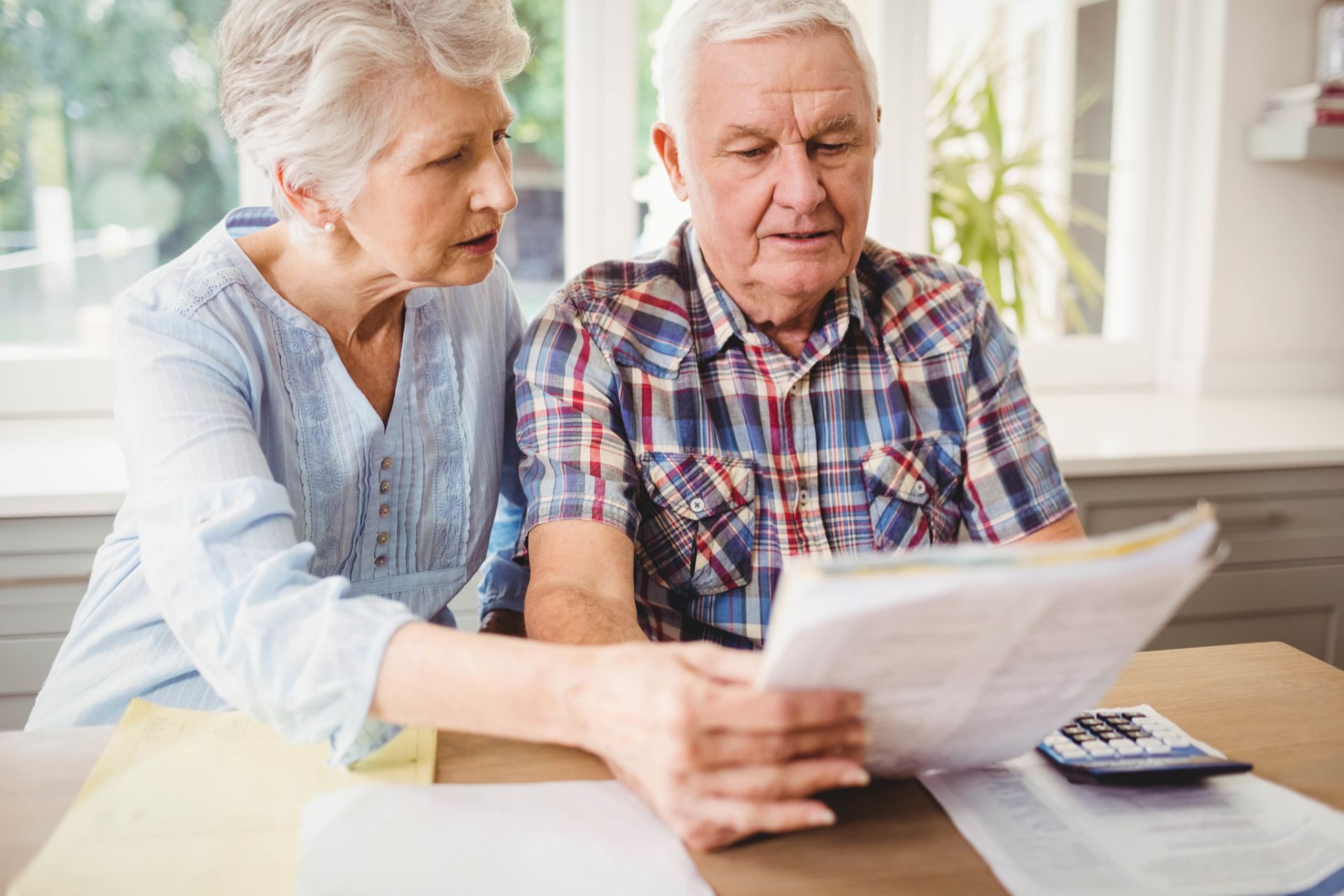 An elderly couple reviews paperwork together, looking concerned; interior setting, with a calculator.