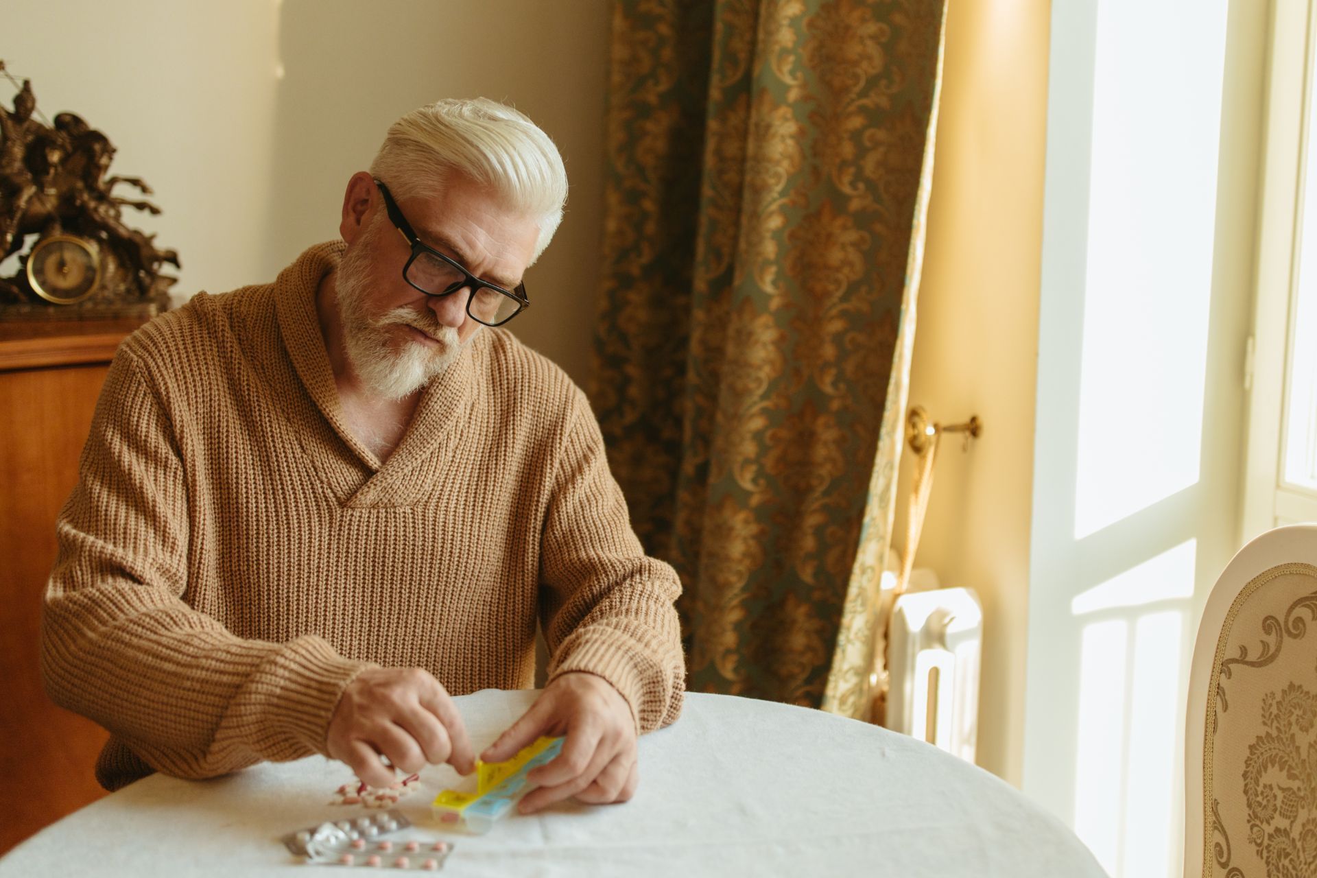 An older man with glasses sorts pills on a table indoors near a window.