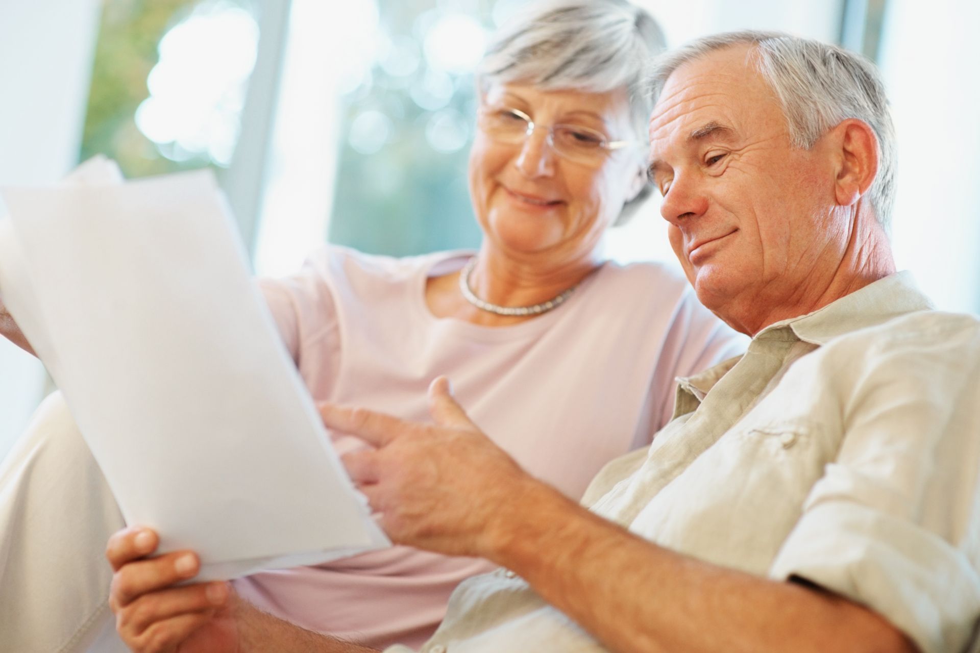 Elderly couple looking at paperwork, smiling. Indoors, neutral colors.