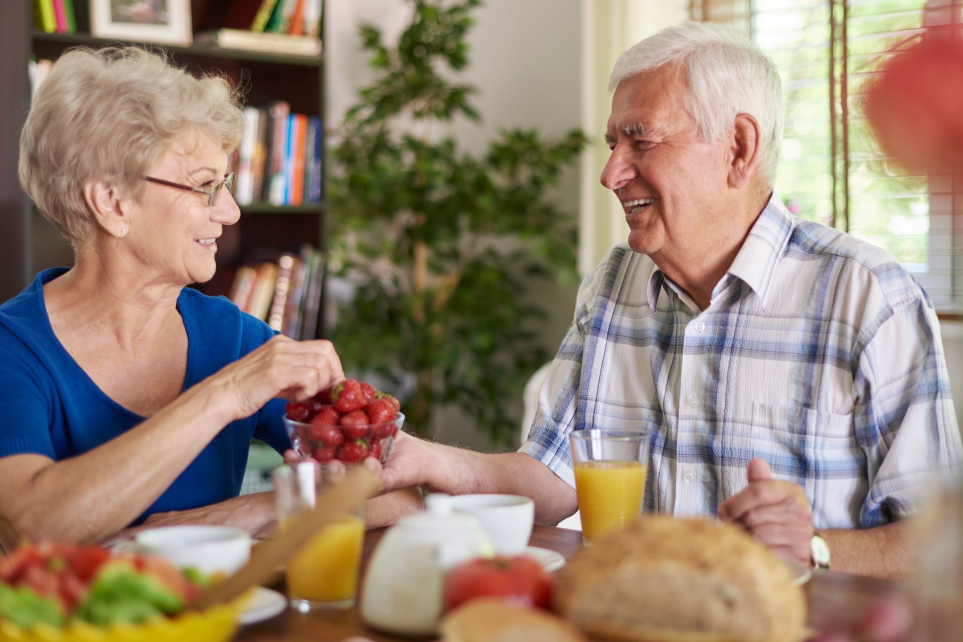 Elderly couple sharing grapes at a breakfast table; smiling, sunny setting.