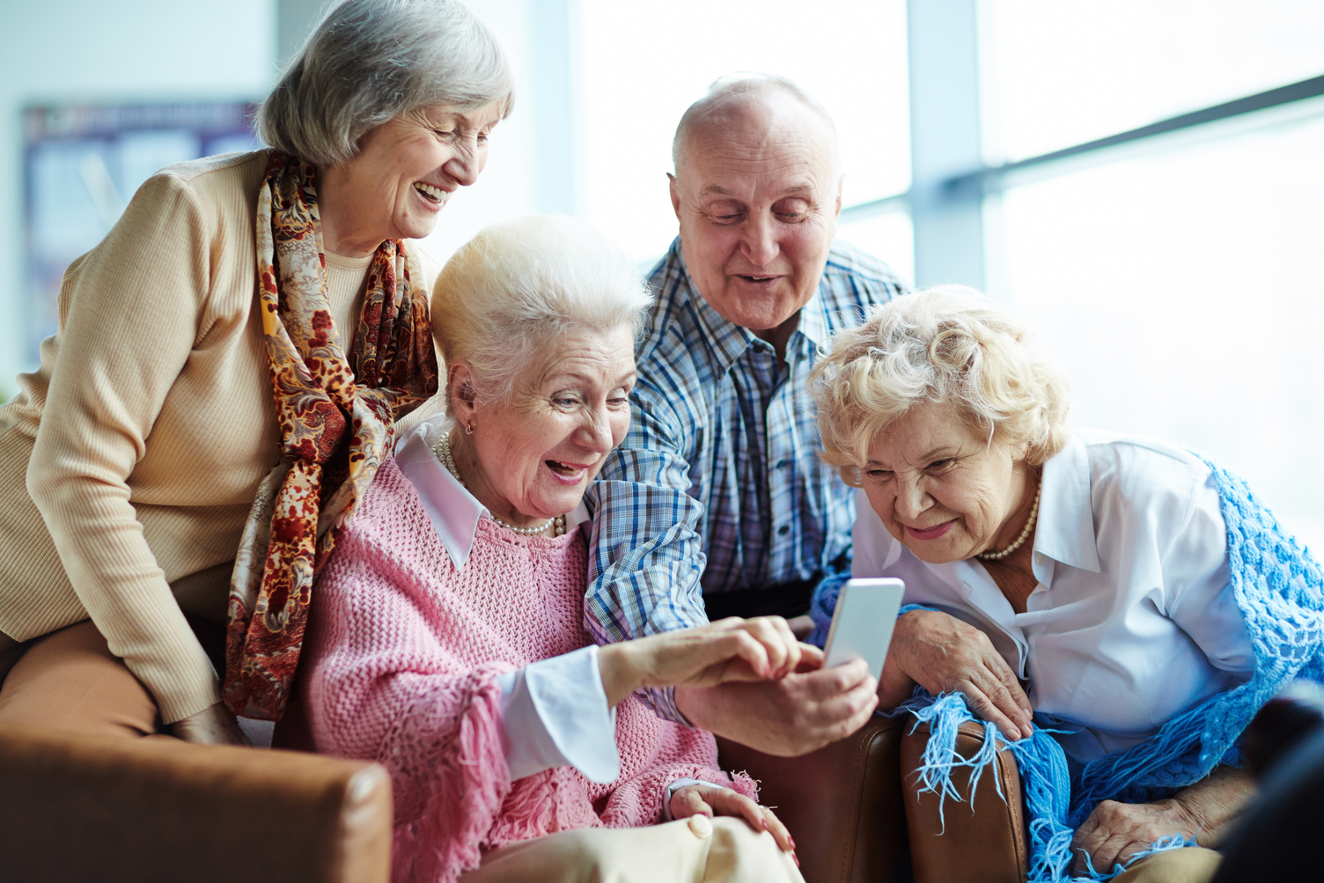 Four older adults smiling, looking at a smartphone together, possibly a care home.