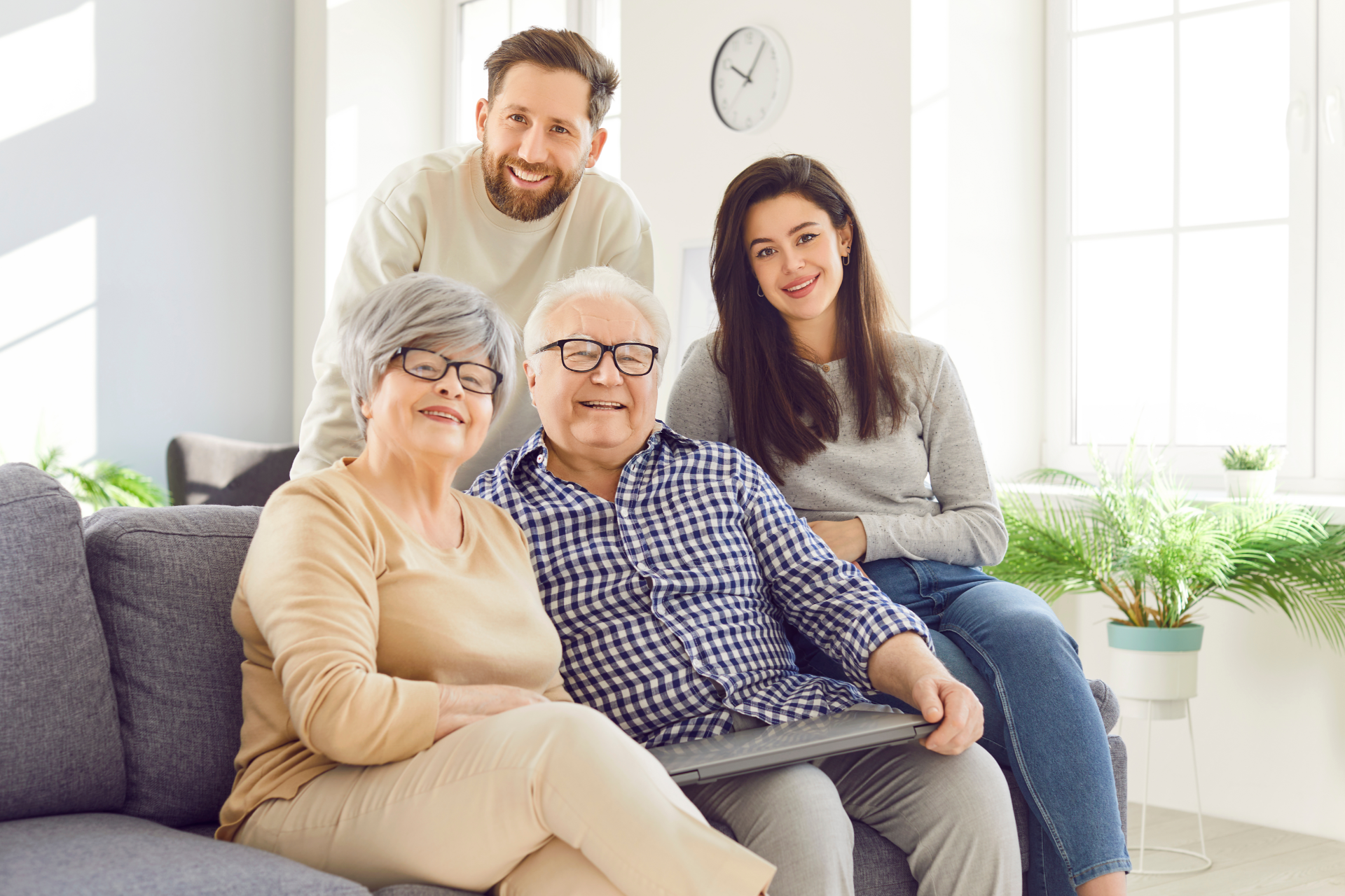 Family portrait: two seniors, two adults, smiling on a couch in a well-lit living room.