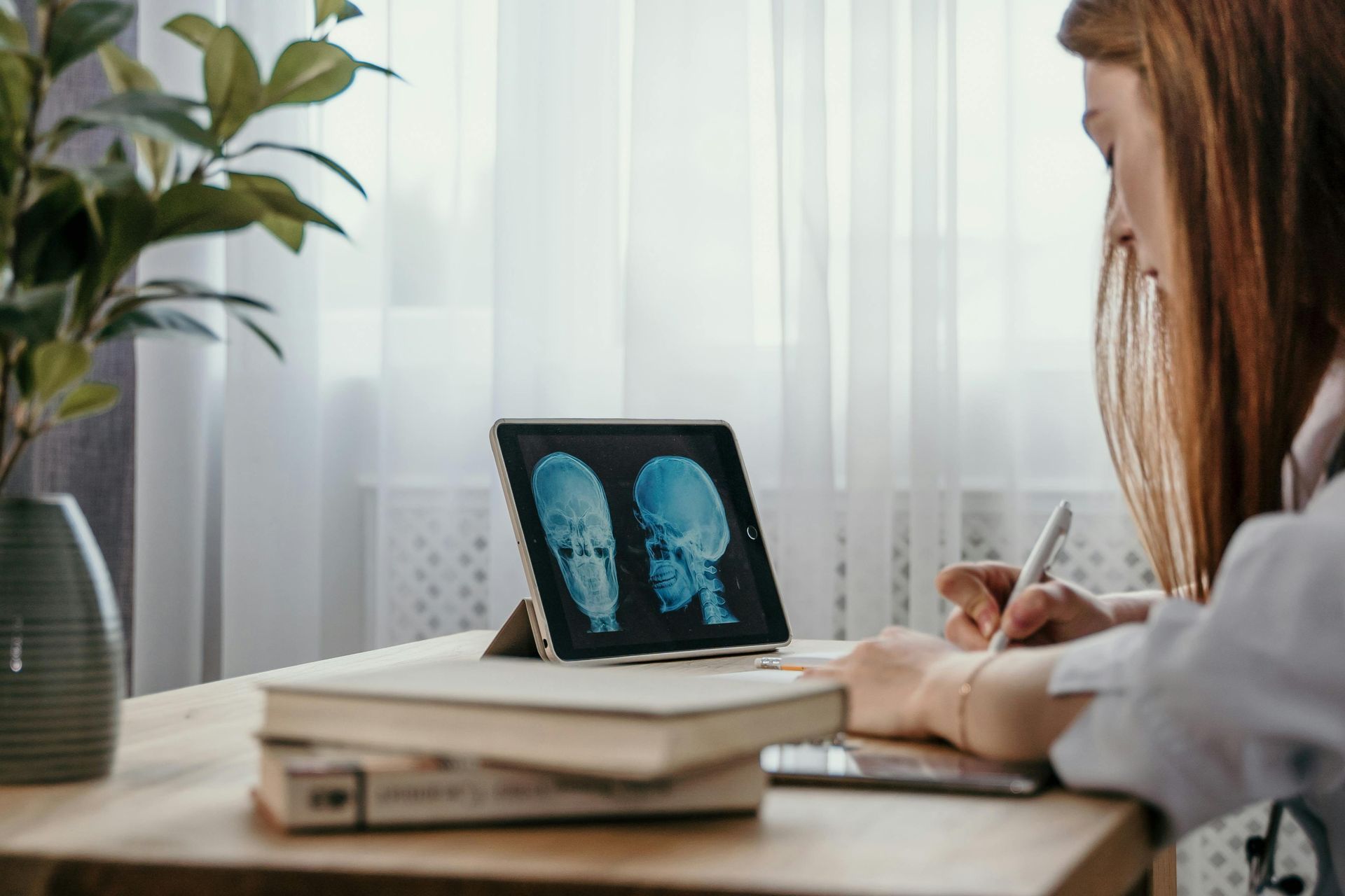 Woman examining brain scans on a tablet, writing notes at a desk near a window and plant.