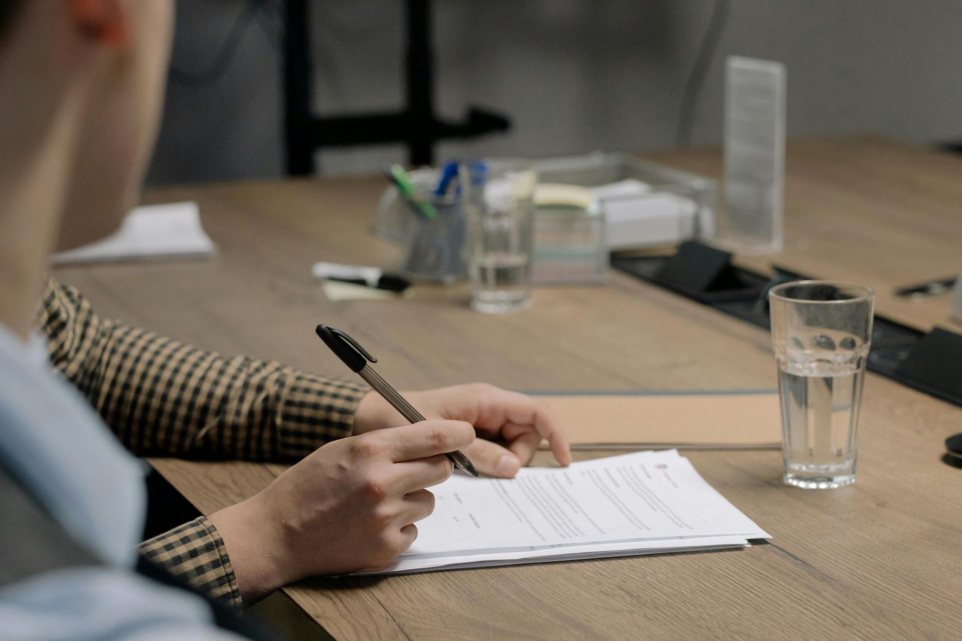 Person in a checkered jacket writing on paper at a wooden table with a glass of water and pens.