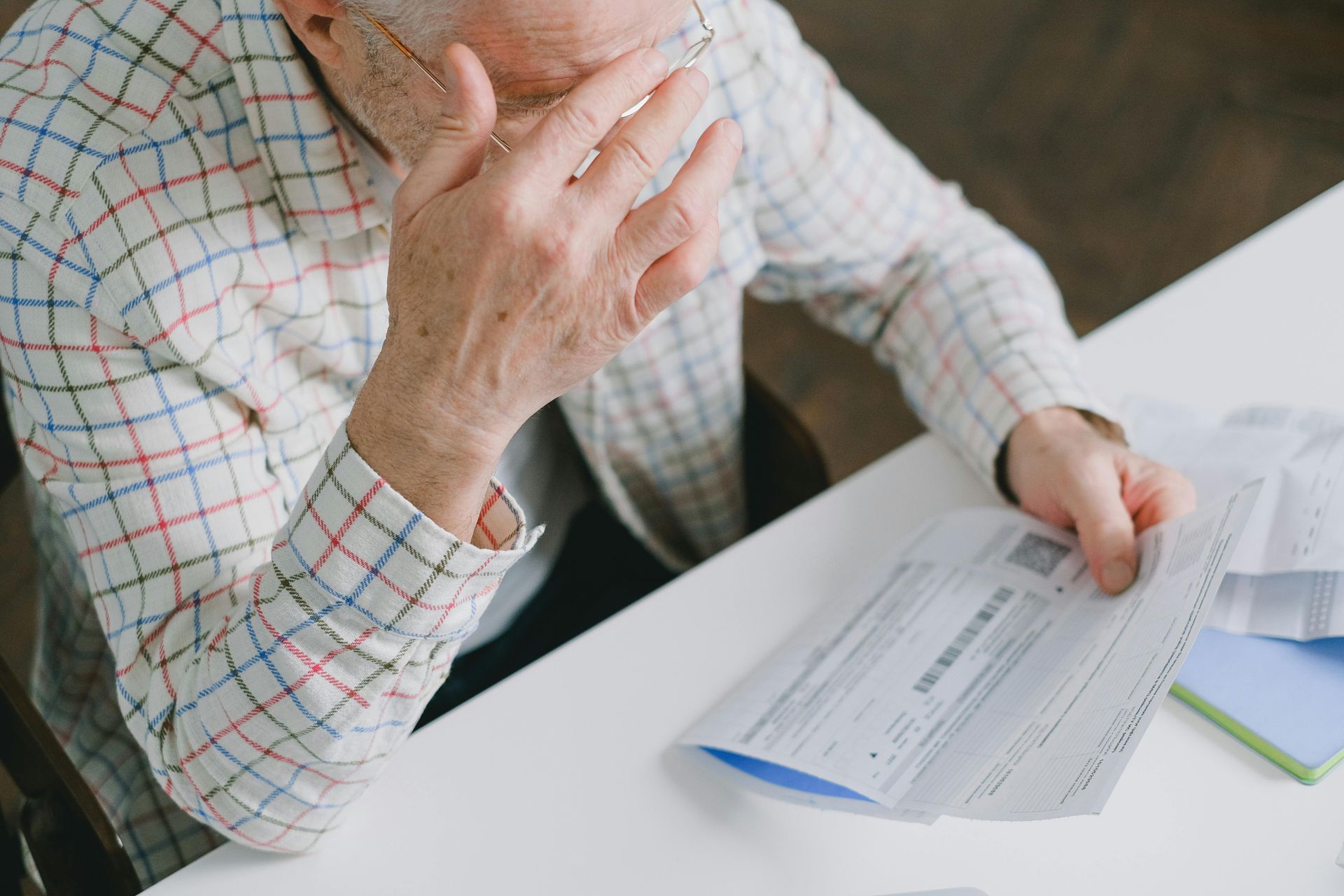 An elderly man stressed, holding bills at a table, covering his face with his hand.