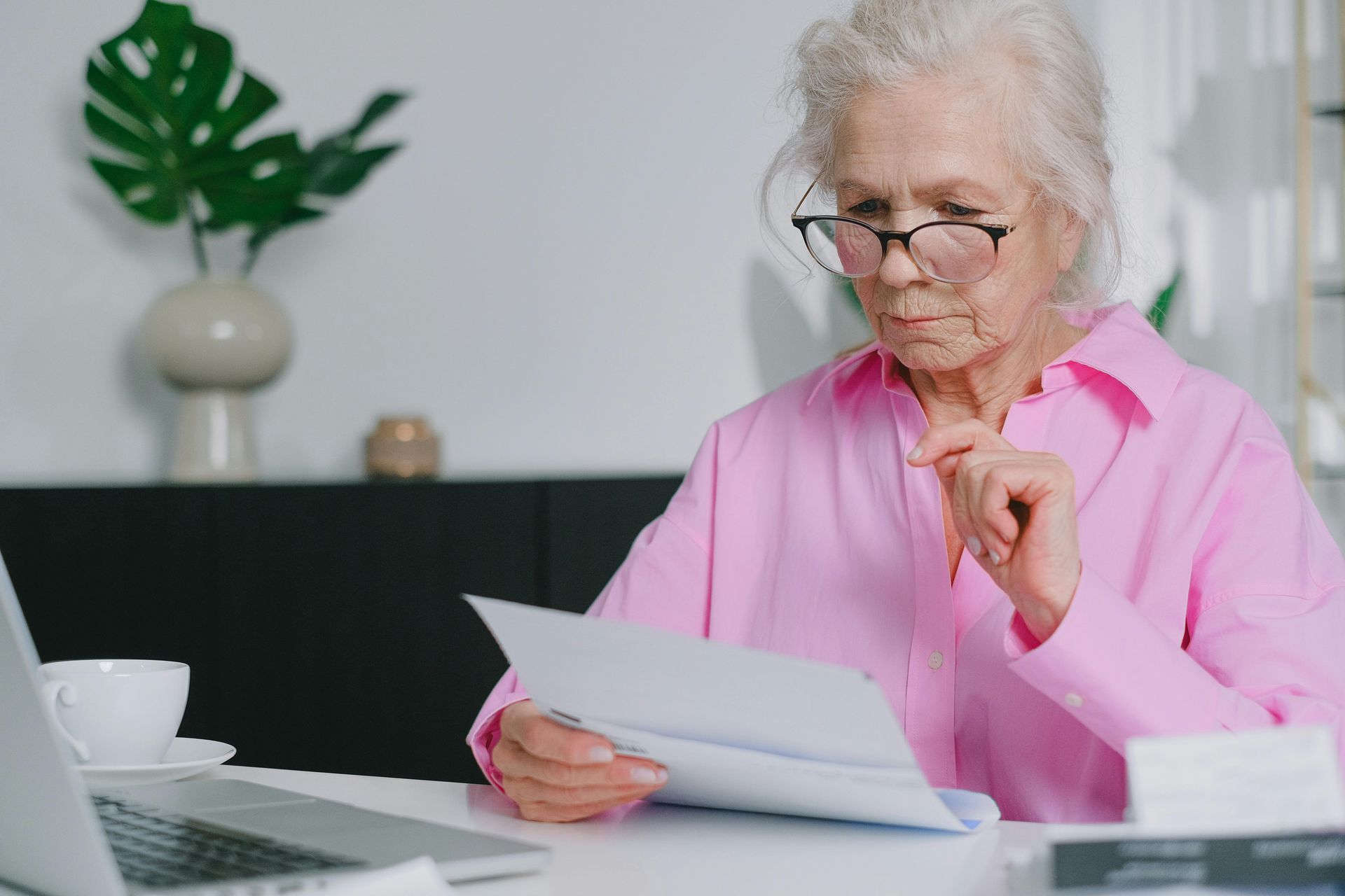 Elderly woman in pink shirt and glasses reviews paperwork at a desk with a laptop.