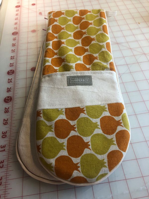 A fabric oven mitt with an orange and green pomegranate pattern, featuring a white pocket with a rectangular brand label.
