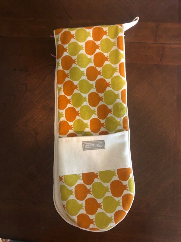 A fabric oven mitt with an orange and green pomegranate pattern, featuring a white pocket with a rectangular brand label.