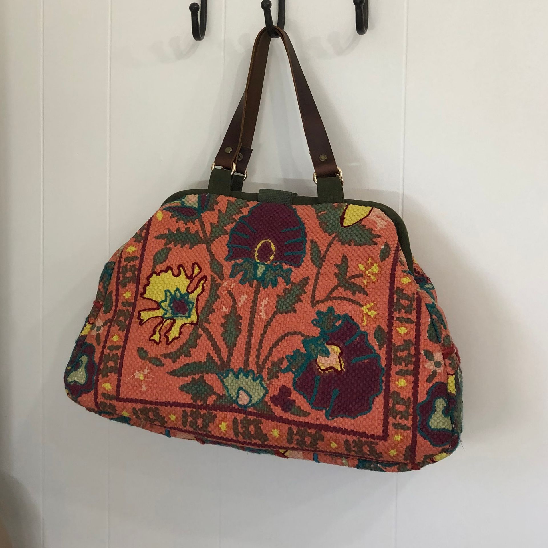 Peach floral tapestry handbag with brown leather handles, hanging on a white wall.