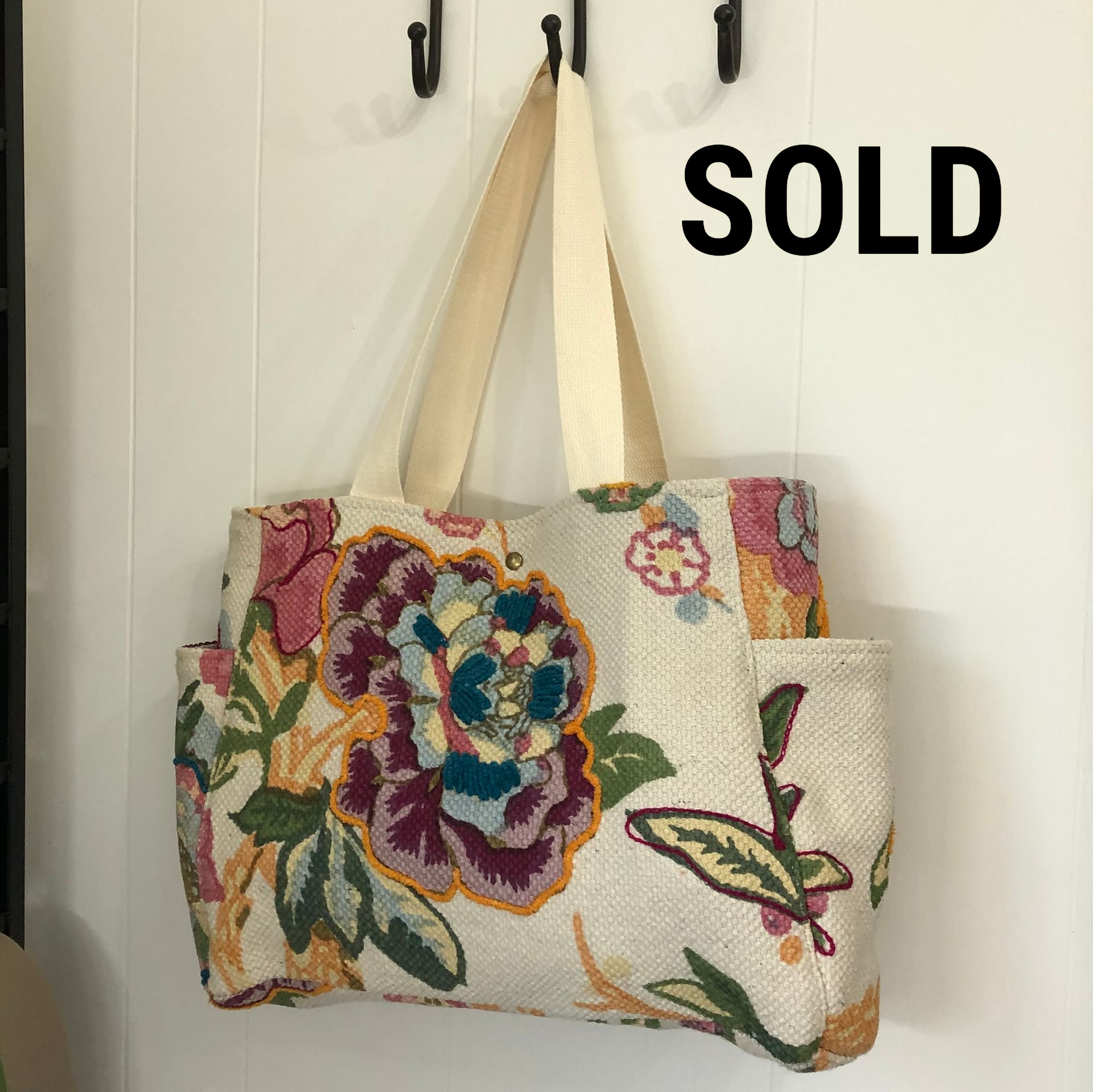 Floral tote bag with cream straps hanging on hooks against a white wall.